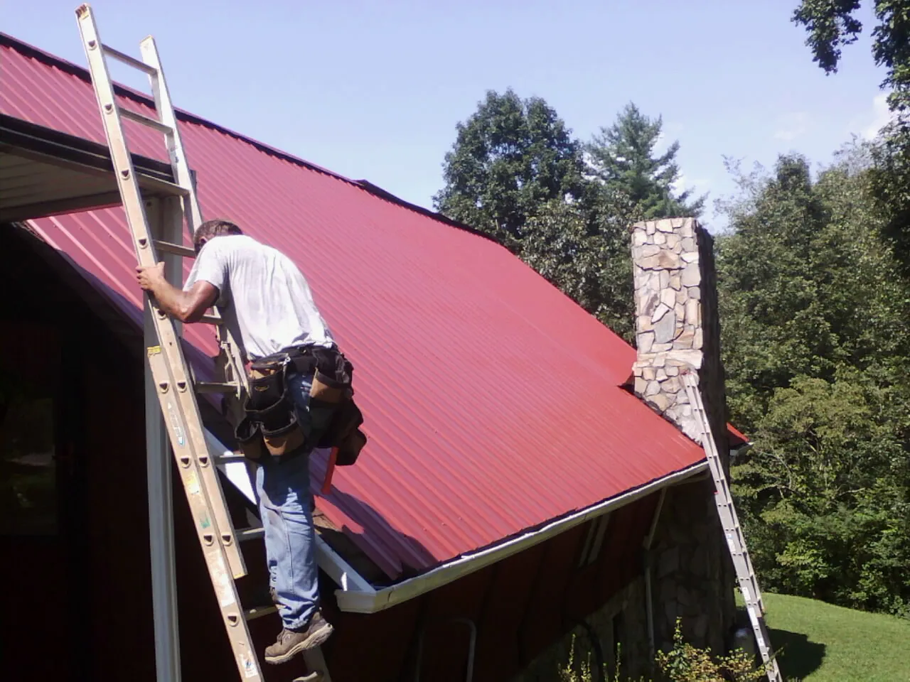 Roofer on a ladder beside a red metal roof and stone chimney in Plymouth